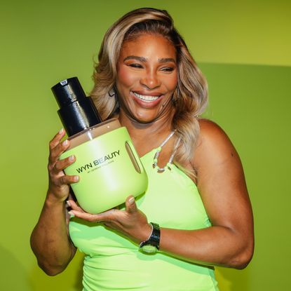 Serena Williams stands in front of a green wall holding a large bottle of wyn beauty foundation
