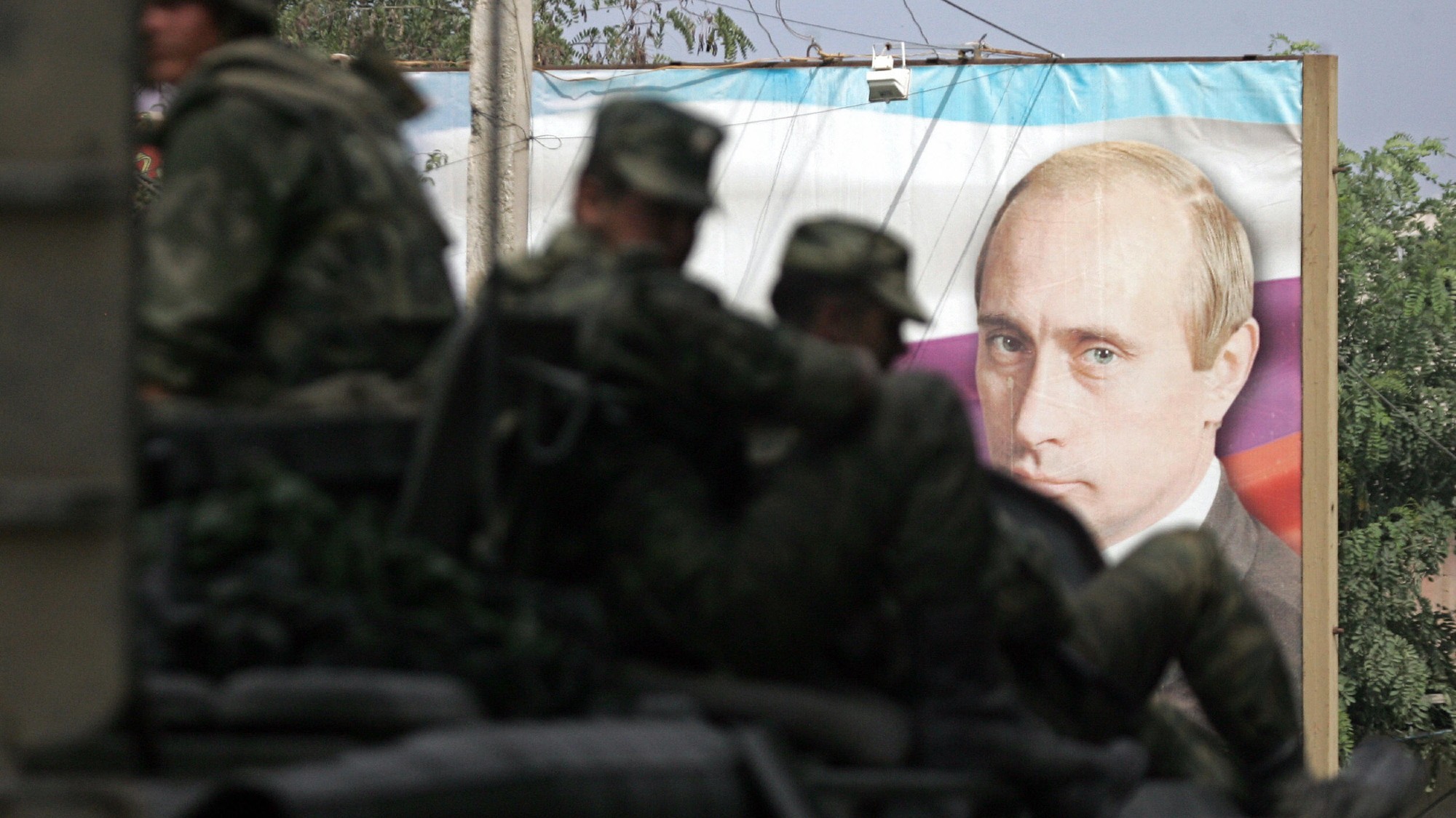 troops aboard an armoured personnel carrier silhouetted against a banner with the face of Vladimir Putin