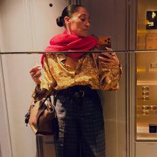 Tracee Ellis-Ross takes a mirror selfie wearing a gold button-down shirt with a pink scarf and checked trousers.