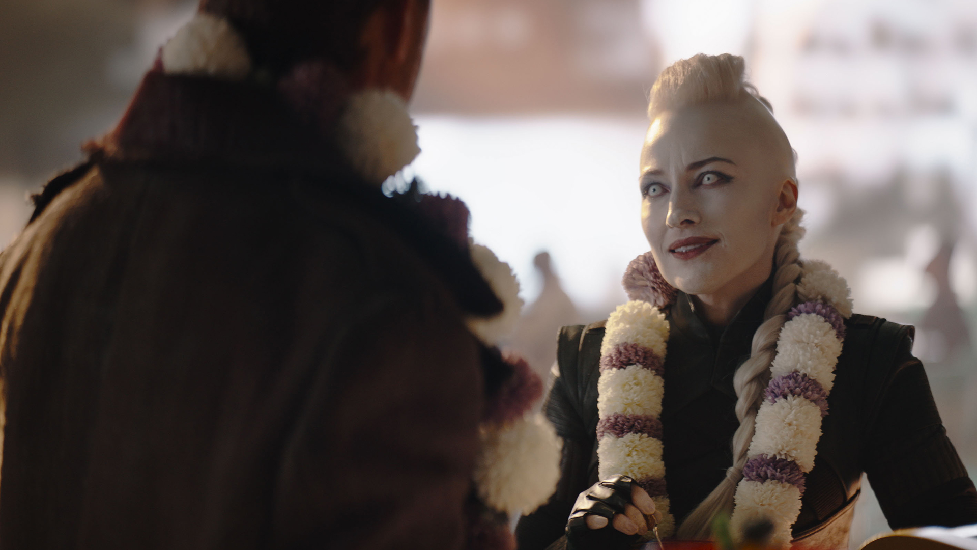 A man with his back to the screen, talking to a woman with pale white skin, white hair, and white eyes.