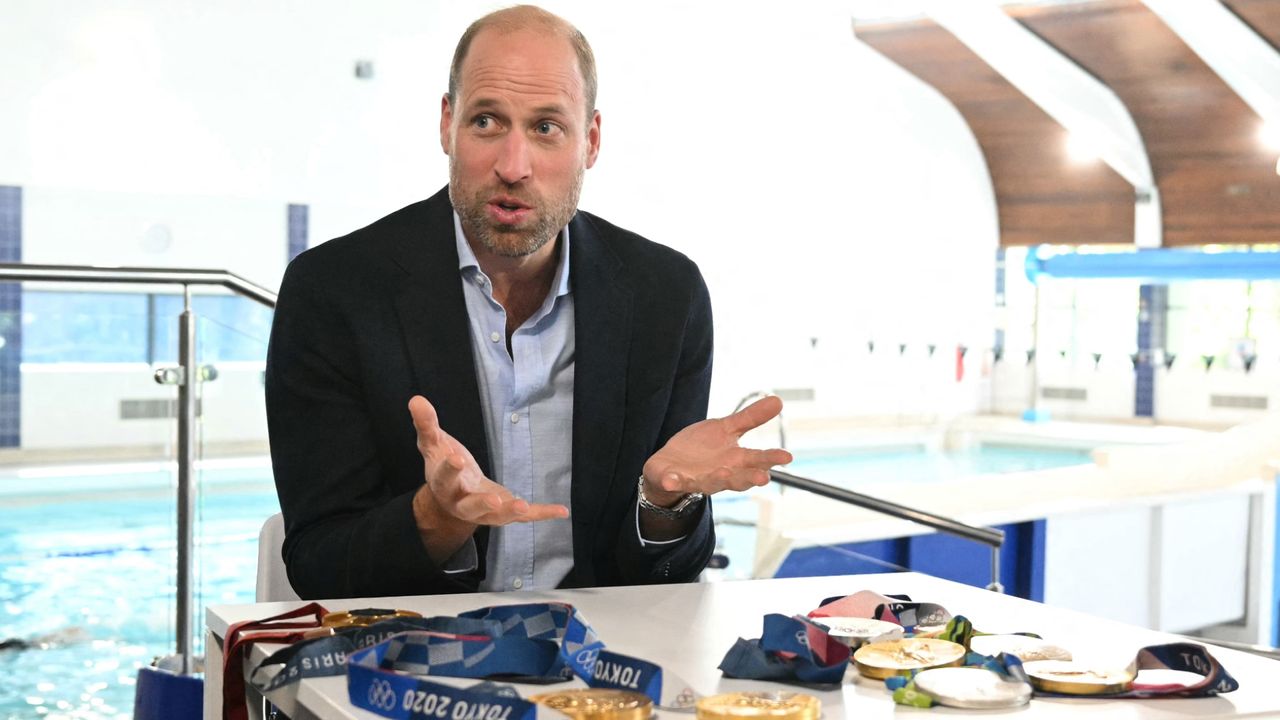 Prince William sitting at a table with Olympic medals in front of a pool