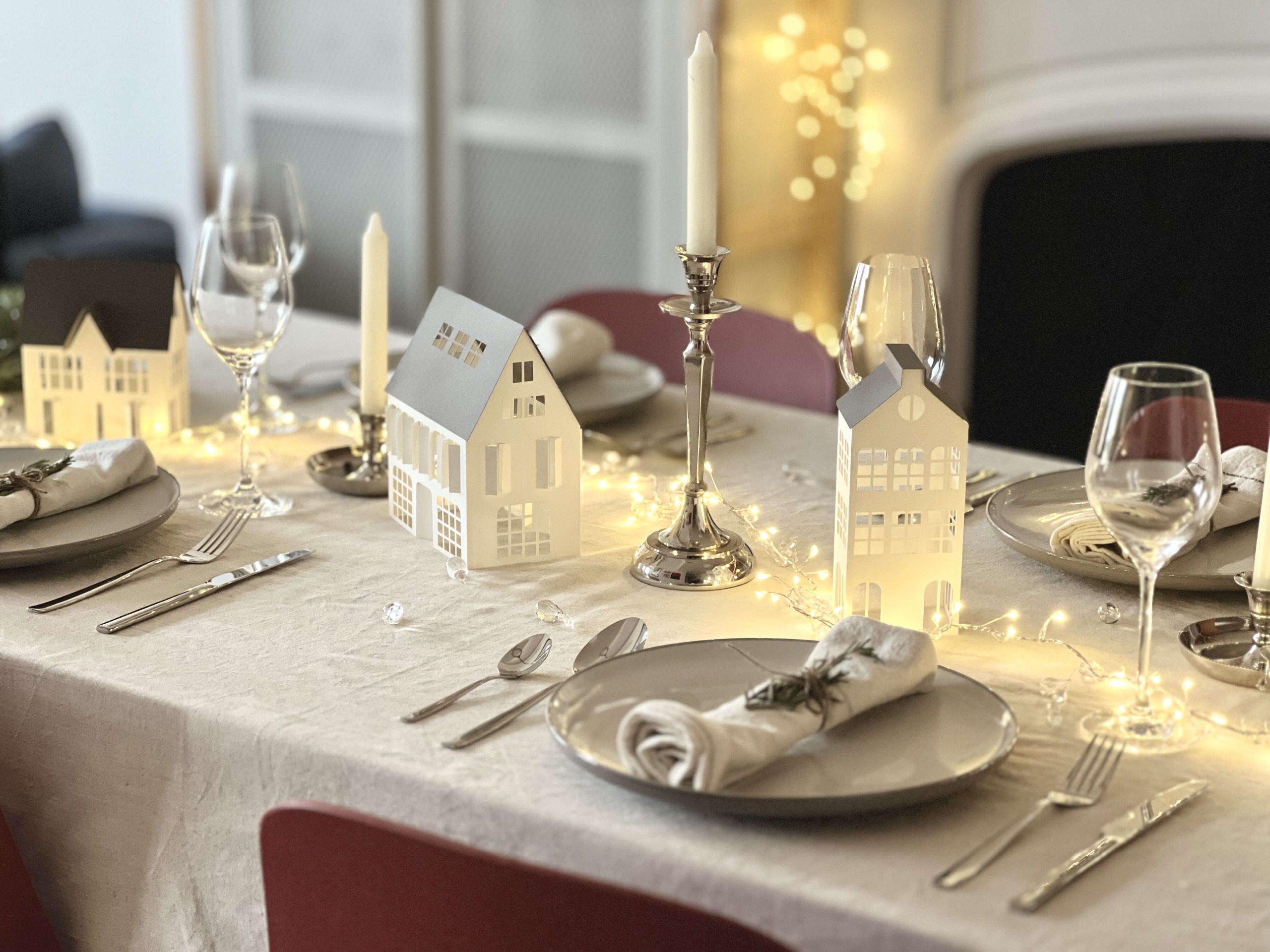 A set dining table with a white tablecloth, napkins on plates, wine glasses and tape candles and string lights down the center with miniature paper christmas houses lining the middle of the table
