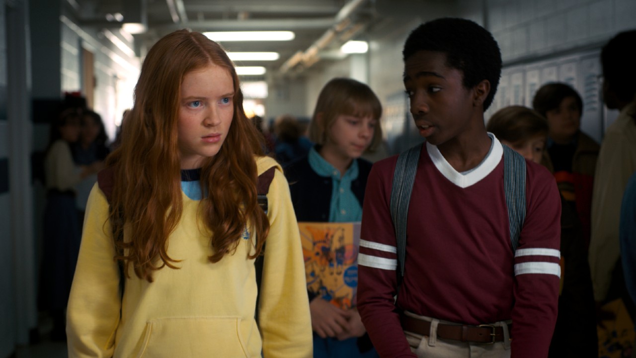 From left to right: Sadie Sink as Max looking forward and Caleb McLaughlin Lucas looking at her. They are standing in a hallway during Season 2 of Stranger Things. 