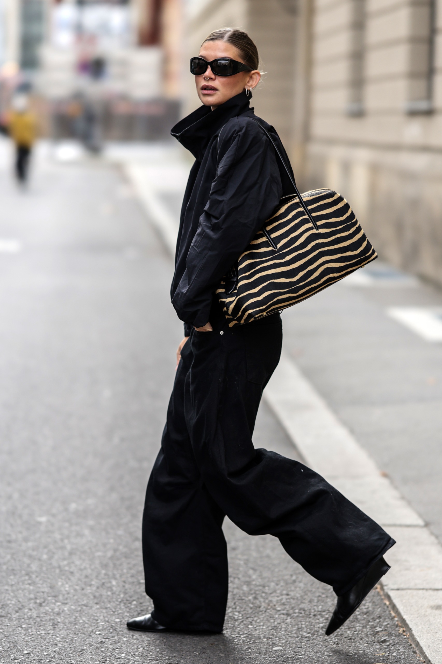 A woman walks outside wearing a black high-neck jacket with black trousers, black shoes and a tiger print bag.