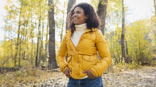 A woman in a yellow jacket walks through a sunlit deciduous forest. She is smiling and her hands are in her pockets. She has shoulder-length curly hair and wears a white high-neck sweater under her coat.