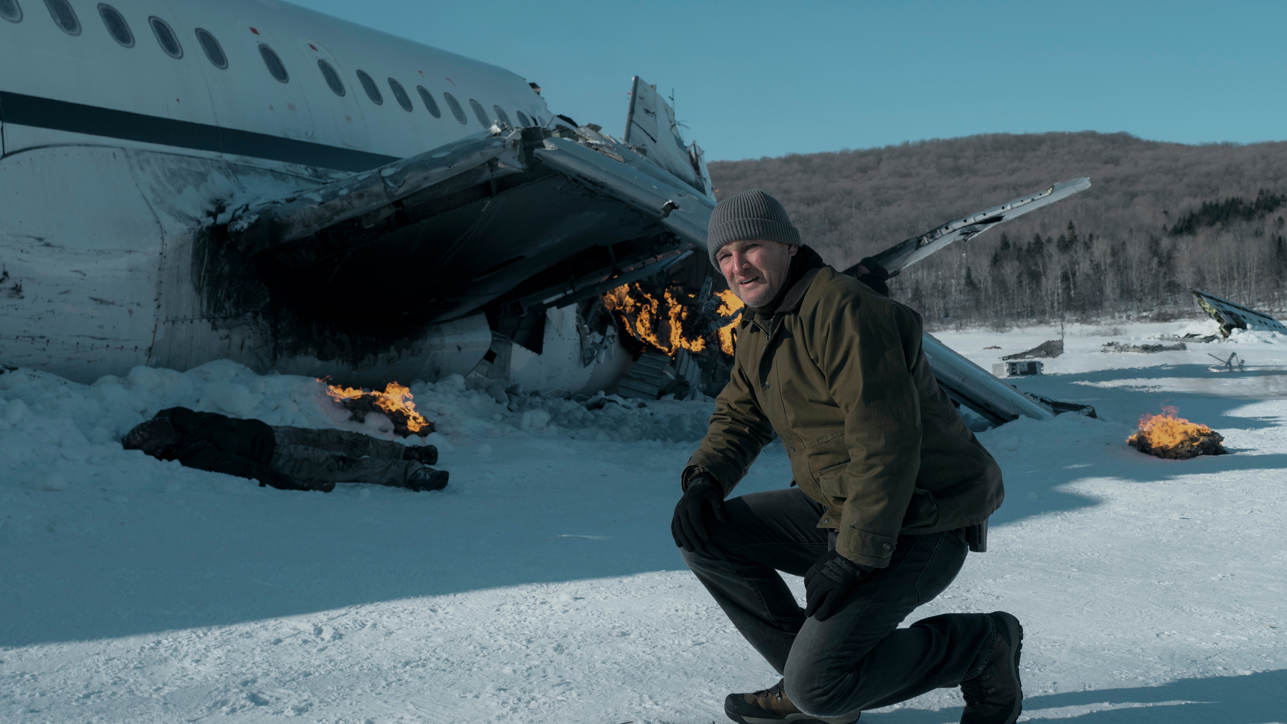 Jason Clarke kneels in front of a burning fuselage