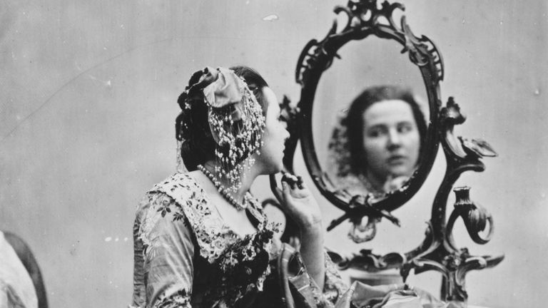 Olden day photograph of lady looking at herself in the mirror wearing a Victorian gown