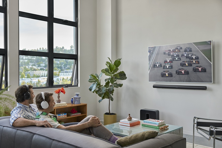 Two models sit watching an F1 race on a TV in a living room, both are wearing the Sonos Ace, thanks to its TV Audio Swap feature.