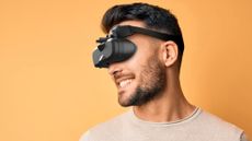 A man with a short beard wearing a MeganeX Superlight mixed reality headset over his eyes. He is in front of a yellow background and is smiling.