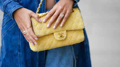 A tight crop of a woman wearing a double denim outfit, holding her hands (with pearlescent nails) over a yellow Chanel bag