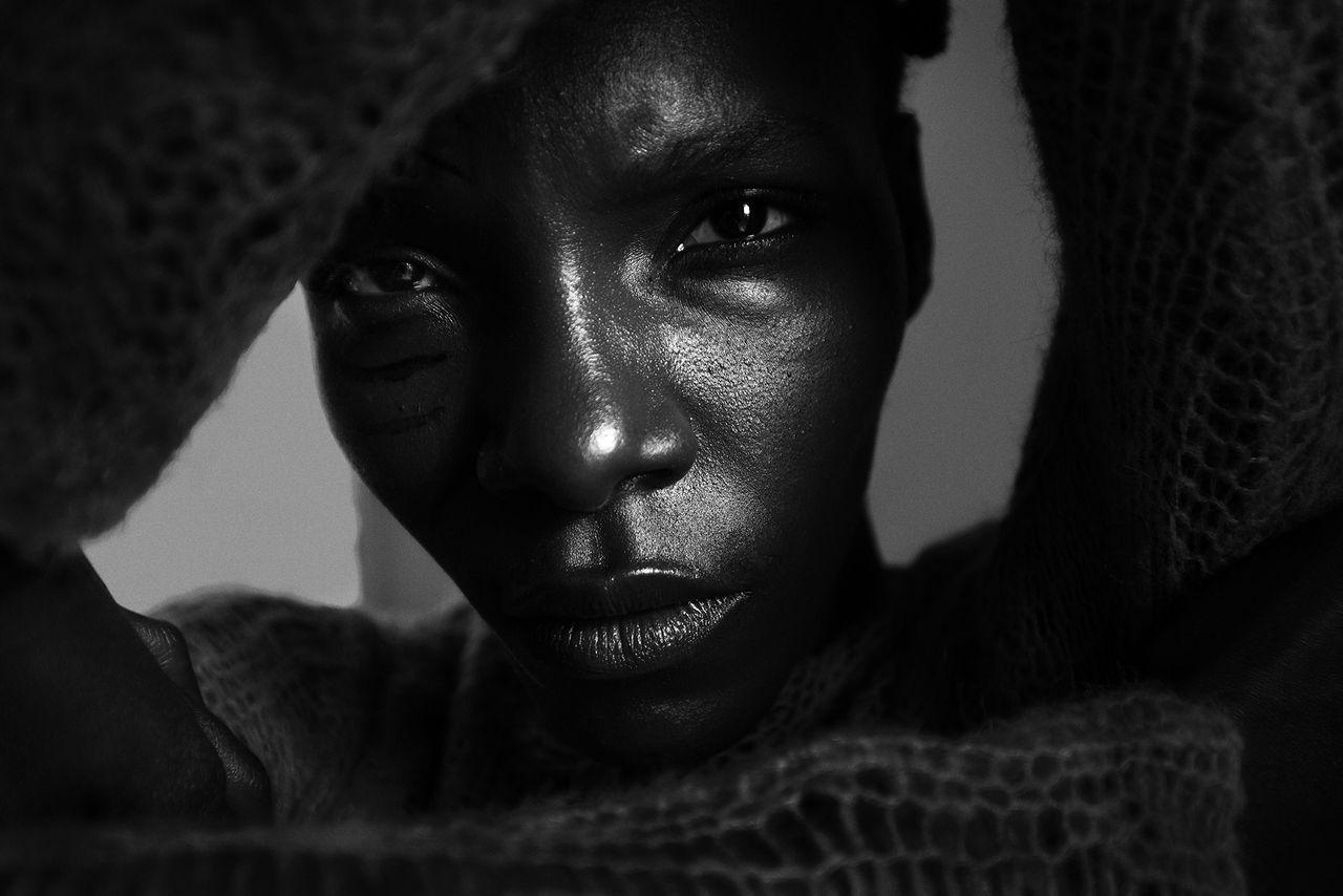 A black and white close-up of a black woman, holding textured fabric in front of her face, featuring an abstract visual composition