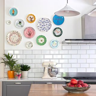 kitchen room with designed plates on white wall and white tiles wall plants pot on counter and fruits in bowl