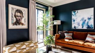 Close-up of a navy living room with a tan velvet sofa, colourful cushions, large blue abstract artwork, tall palm plant and a gold floor lamp against a dark wall.