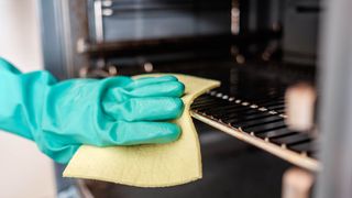 Someone cleaning an oven rack inside an oven with a cloth while wearing gloves