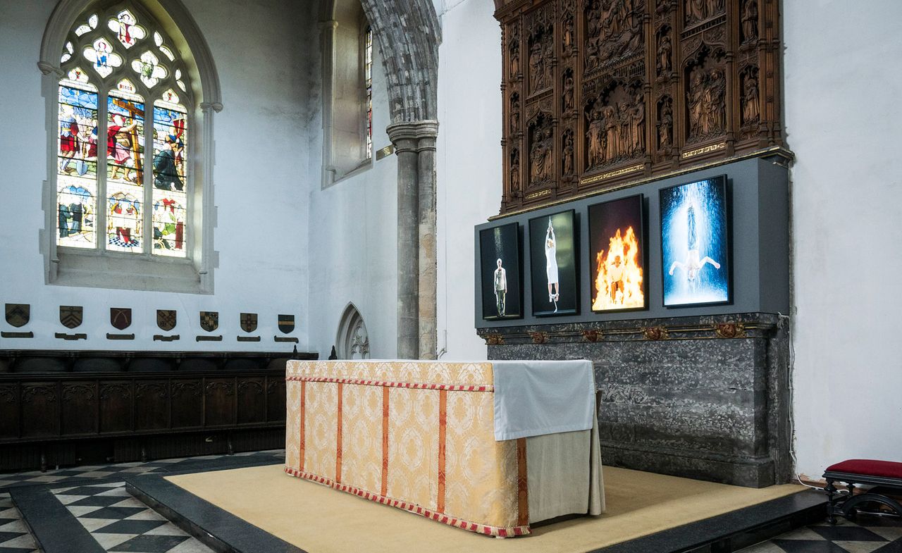 Auckland Castle interior, white walls, arched stained glass window, decorated cloth covered alter on a black platform, black and white diamond design stone floor, stone pillar, red seat, row of small wooden shields on left wall under window, stone and wood centre piece on right wall four pieces of art work incorporated 