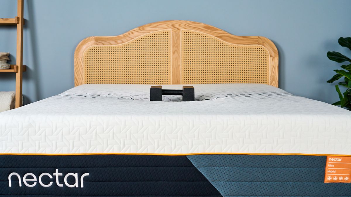 The Nectar Ultra Hybrid Mattress on a bed frame in a bedroom, a kettlebell balanced on top to show pressure relief. The weight has sunk so far into the mattress only the handle shows