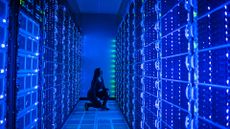 Data center server room glowing with bright blue and green colors with female employee kneeling inspecting server rack.