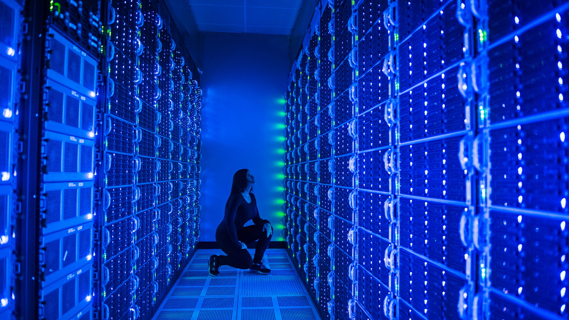 Data center server room glowing with bright blue and green colors with female employee kneeling inspecting server rack. 