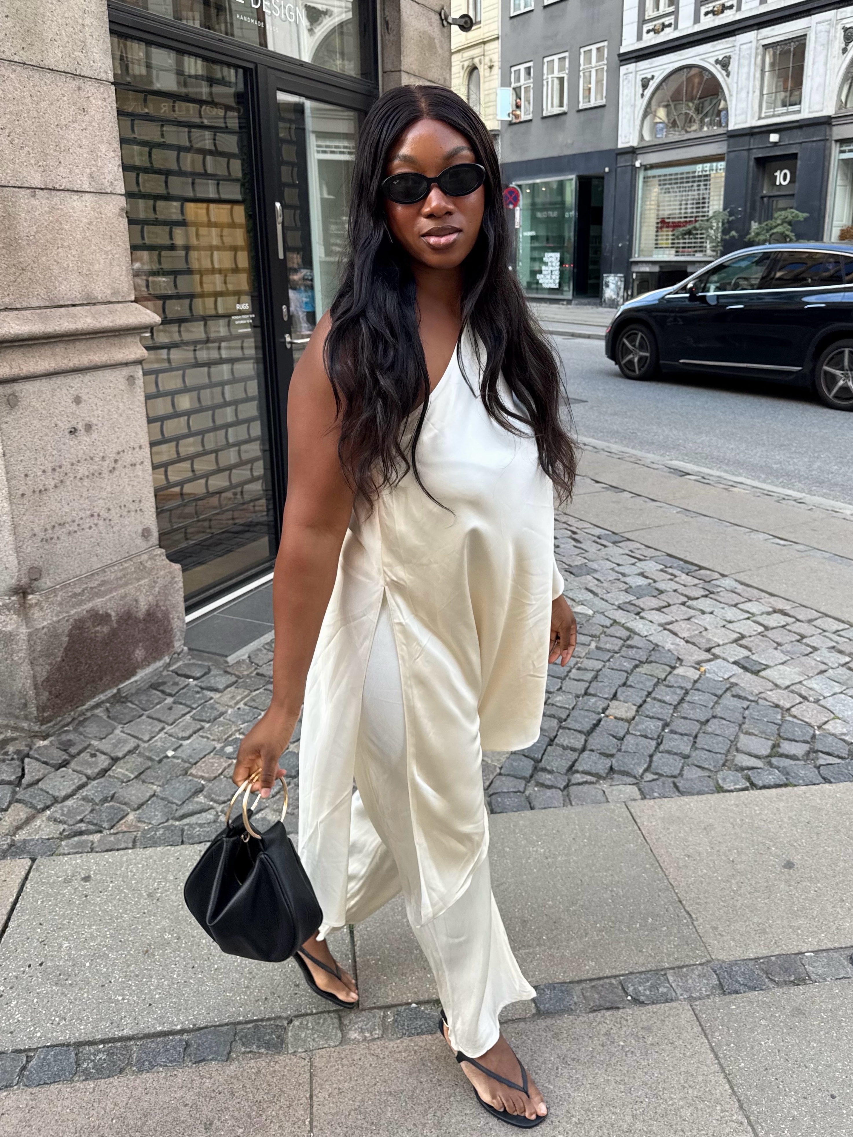 An image of an influencer wearing white satin trousers and a matching white one-shoulder satin top and black heeled sandals carrying ring-handled tote