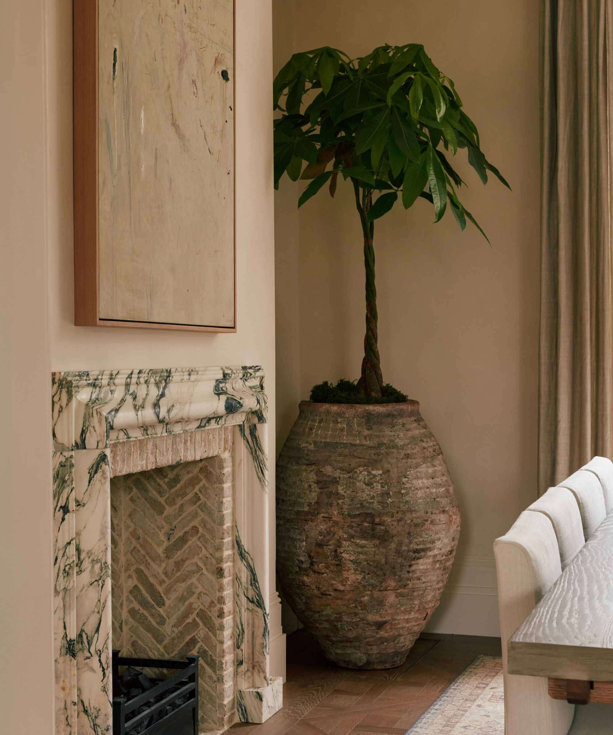 Corner of living room that shows a large plant in a rounded stone planter, marble fireplace, artwork hung above, corner of white sofa on left-hand side