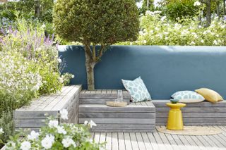 Designing a patio with layered decking, set against a blue wall, yellow and blue accessories and small tree.