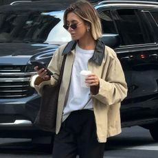 British style influencer Hannah Lewis crosses a street while looking at her phone wearing a tan barn coat, white T-shirt, black pants, and black wire cat-eye sunglasses
