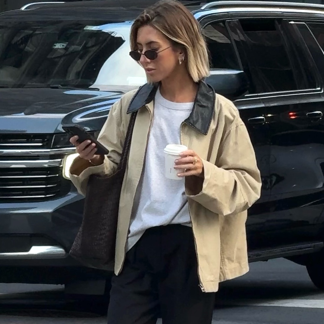 British style influencer Hannah Lewis crosses a street while looking at her phone wearing a tan barn coat, white T-shirt, black pants, and black wire cat-eye sunglasses