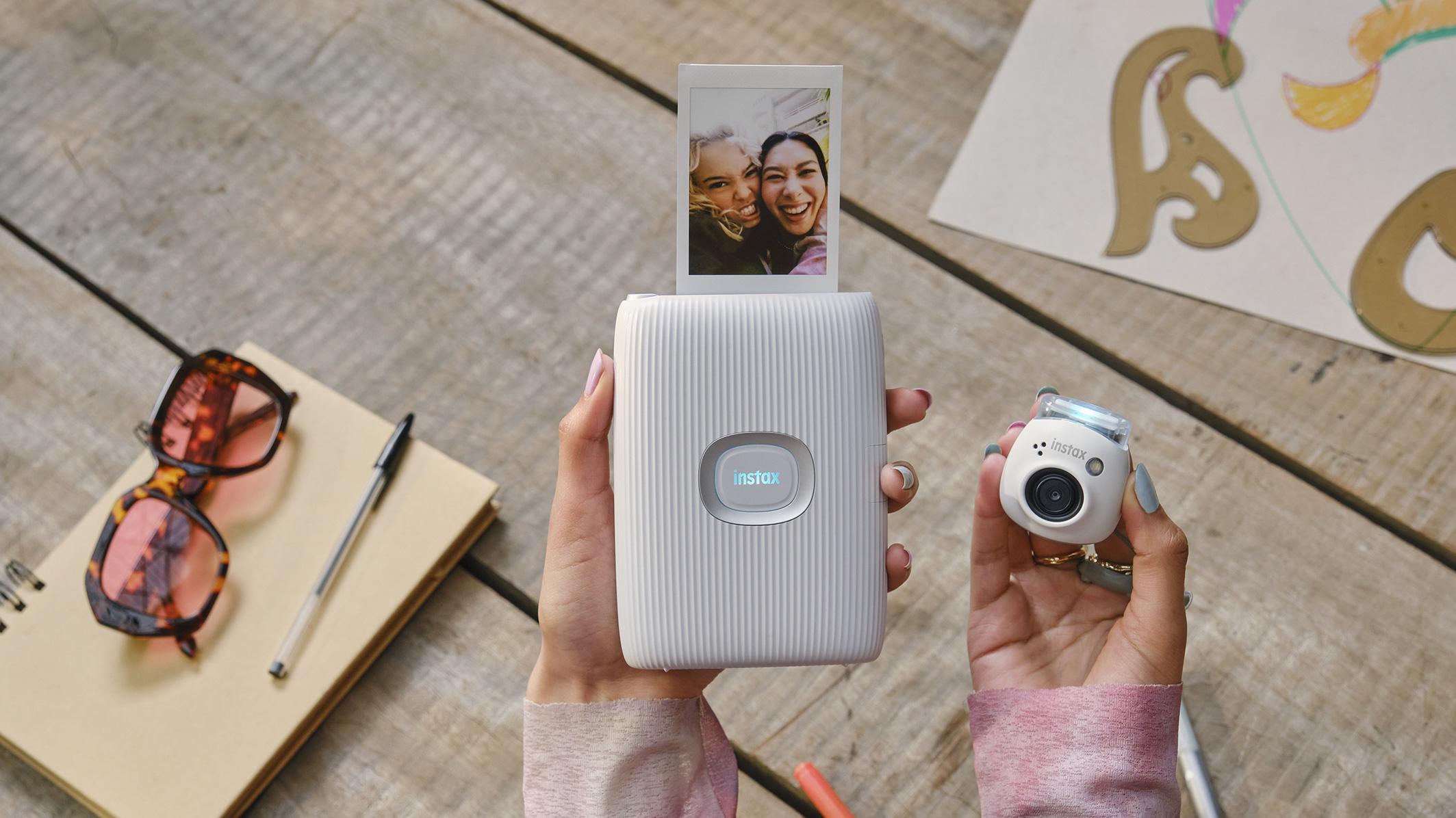 Fujifilm Instax Pal in the hand alongside a Instax Link printer with an instant film churned out