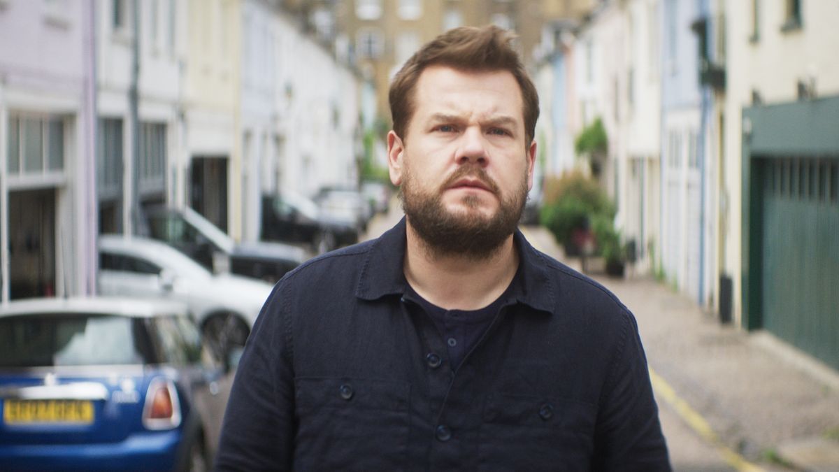 James Corden in a dark top looks pensive standing in a street in Mammals