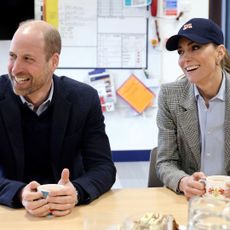 Princess Kate smiling in a baseball cap sitting next to Prince William at a table