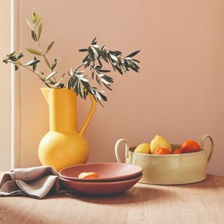 A wooden dining table against a pink wall with a yellow jug and and a bowl of citrus fruit on top