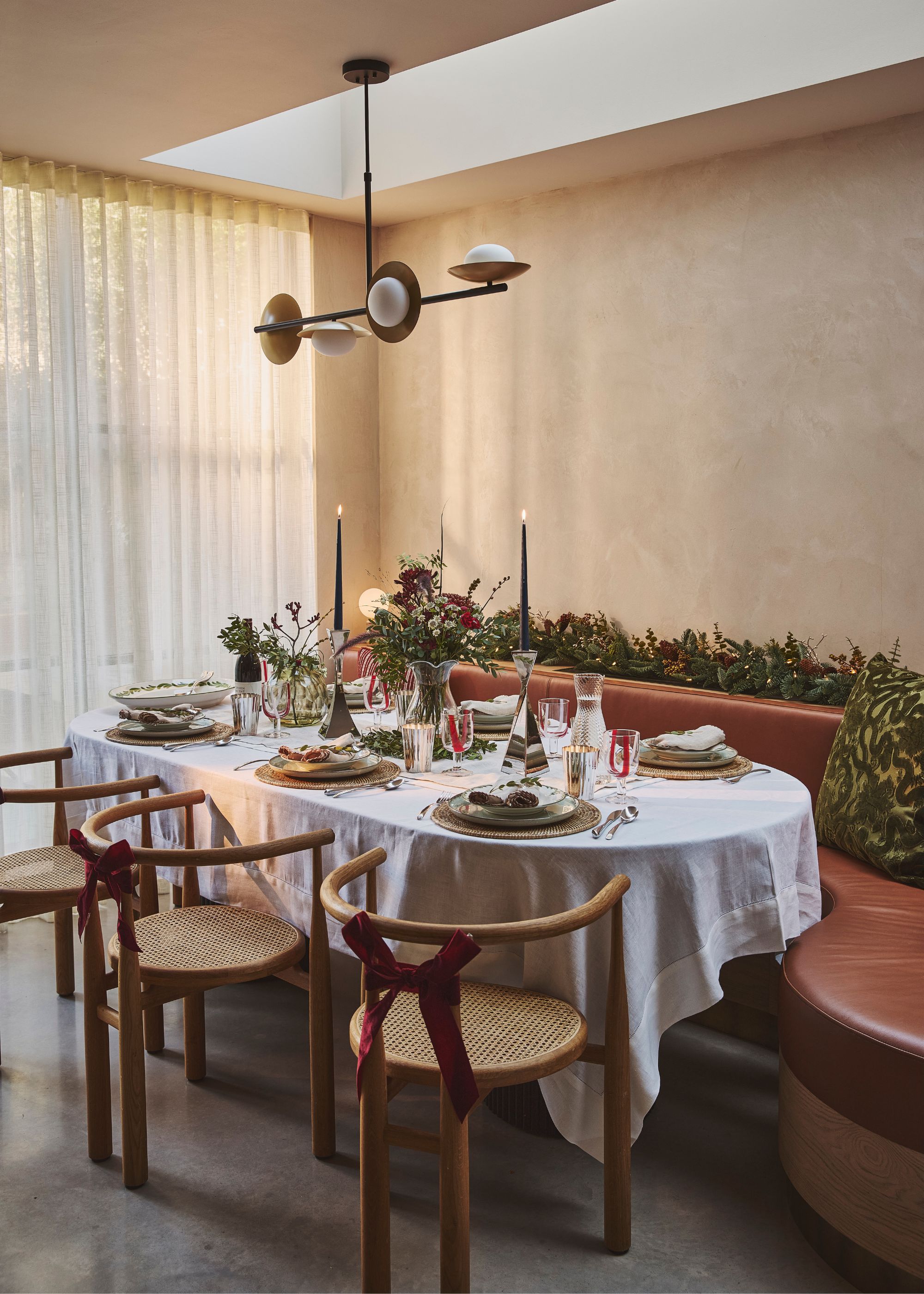 A modern dining room with a festive tablescape with a white tablecloth and long candles, wooden dining chairs, and banquette seating.