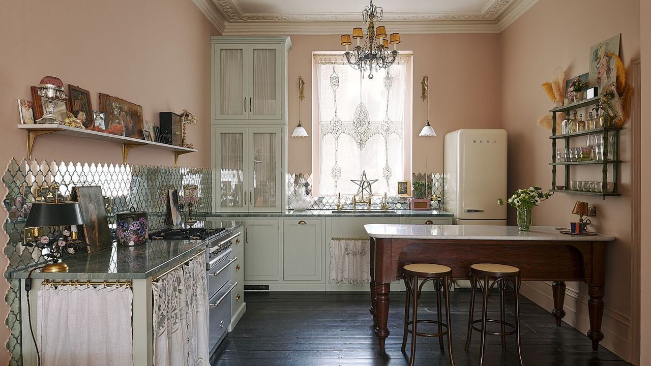 Pastel kitchen with soft pink walls, light pastel green cabinets, retro cream SMEG fridge, dark mahogany dining table, silver tiles, dark wooden floors and shelves packed with mementos, along with crown molding on ceiling and vintage lace curtains and lights