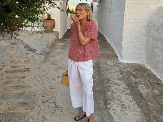 Fashion influencer Lucy Williams on summer vacation in Europe wears drawstring white pants, a breezy short-sleeve blouse, and flat strappy sandals.