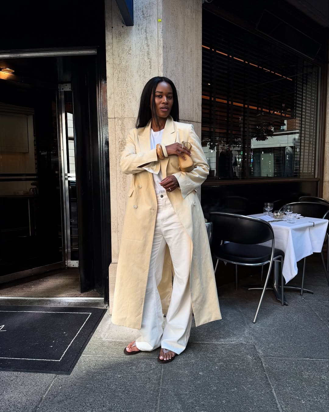 Influencer Marilyn wears a pair of white denim jeans, a white crewneck tank, and a butter yellow long coat.
