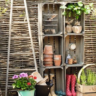 Woven willow branch garden fence, French ladder, vintage fruit boxes and crates as open storage shelves, storing, flowerpots, wellies, turquoise enamel bucket, plants, wire baskets, recycled rubber tyre