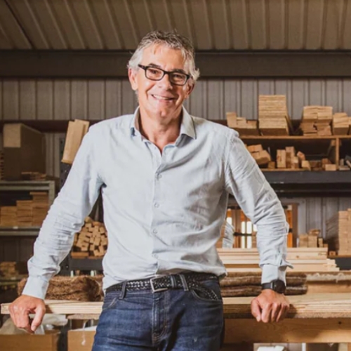 Image of a man in a long-sleeve, light blue button-down and jeans. He is standing in a wood workshop.