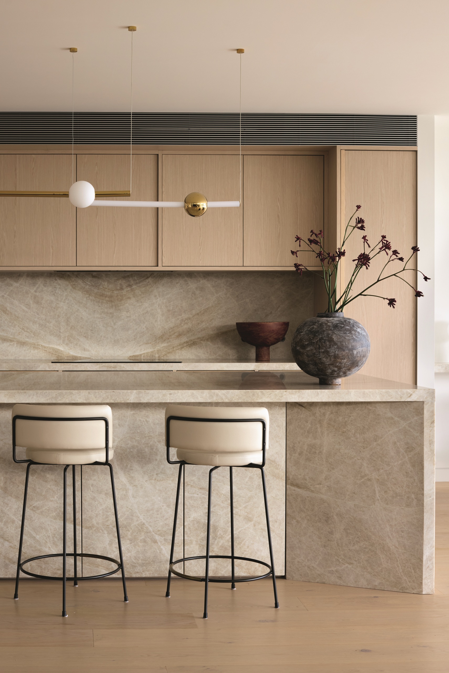 neutral kit chen with angular stone island with black-and-white bar stool, light timber cabinetry, and dark round vase with branches