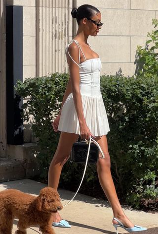 a woman's outfit showing what to wear in 80-degree weather with a white minidress styled with blue denim slingback pumps, a black handbag, and black sunglasses