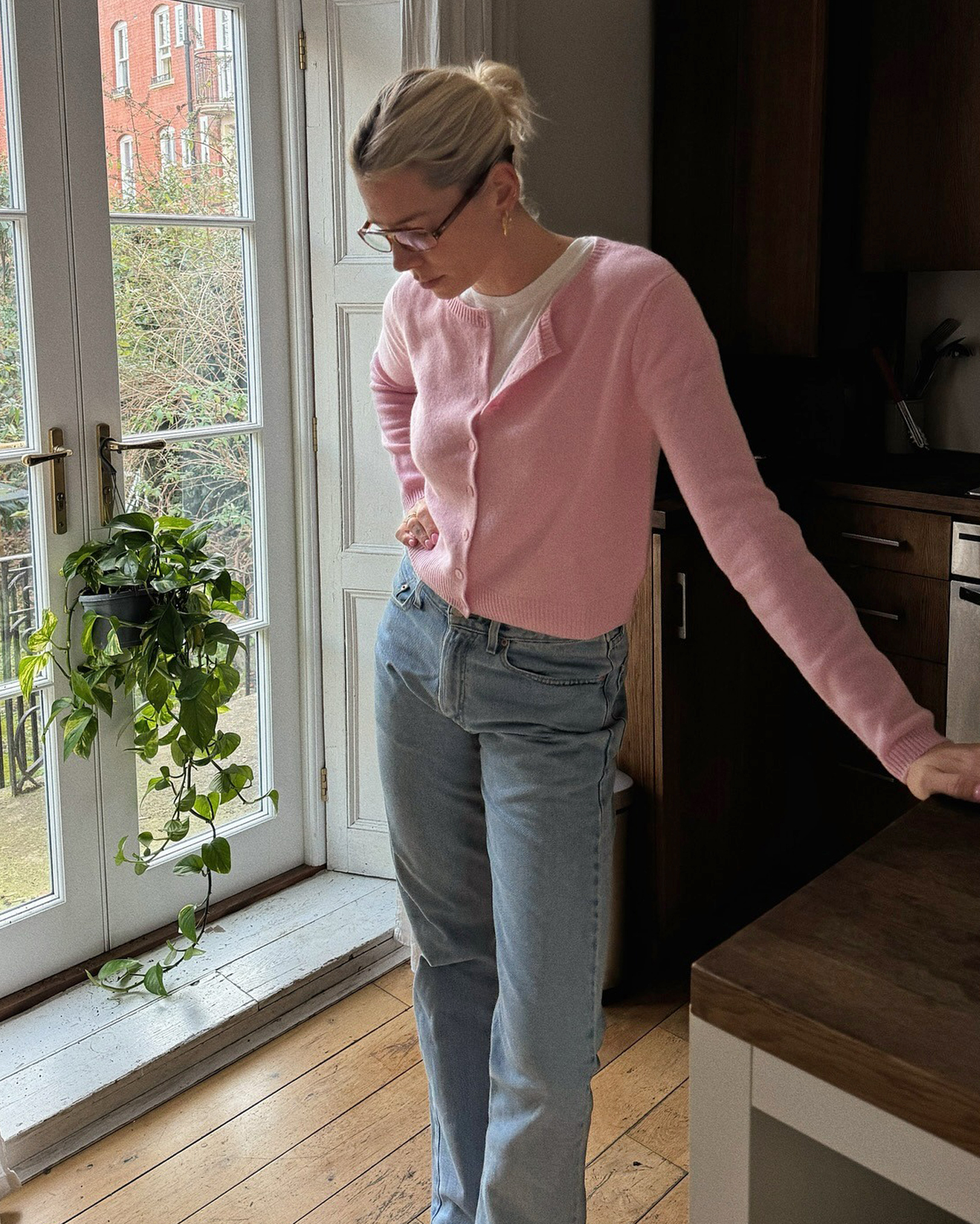 British style influencer Lindsey Holland poses in her kitchen wearing eyeglasses, a pink button-down cardigan layered over a white T-shirt, and jeans.
