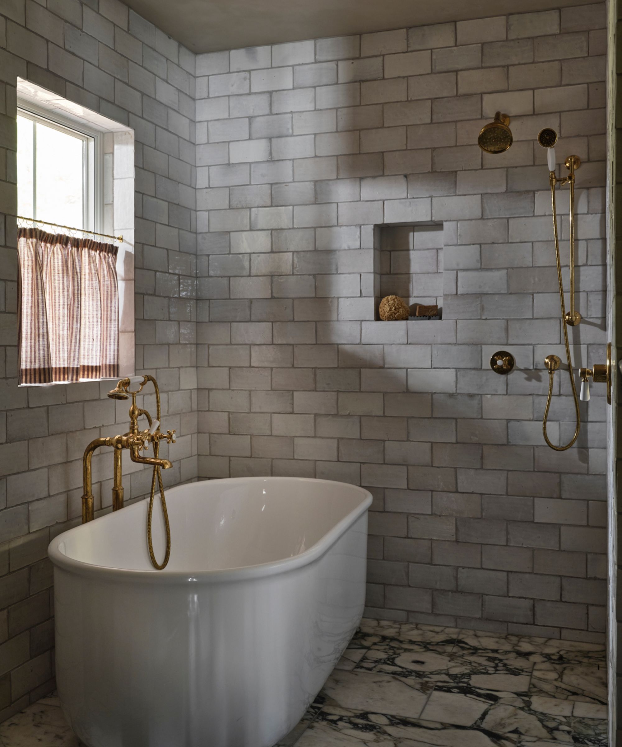 an earthy muted wet room bathroom with marble floor tiles, a free standing tub, subway zellige tiles on the walls and a small niche