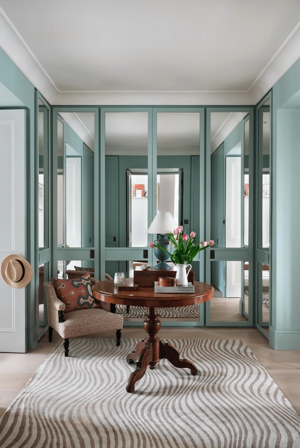 Acquamarine-tinted interior, featuring mirrored walls, a rug with wavy brown lines on beige background, a central wooden table, an armchair, flowers, and books.