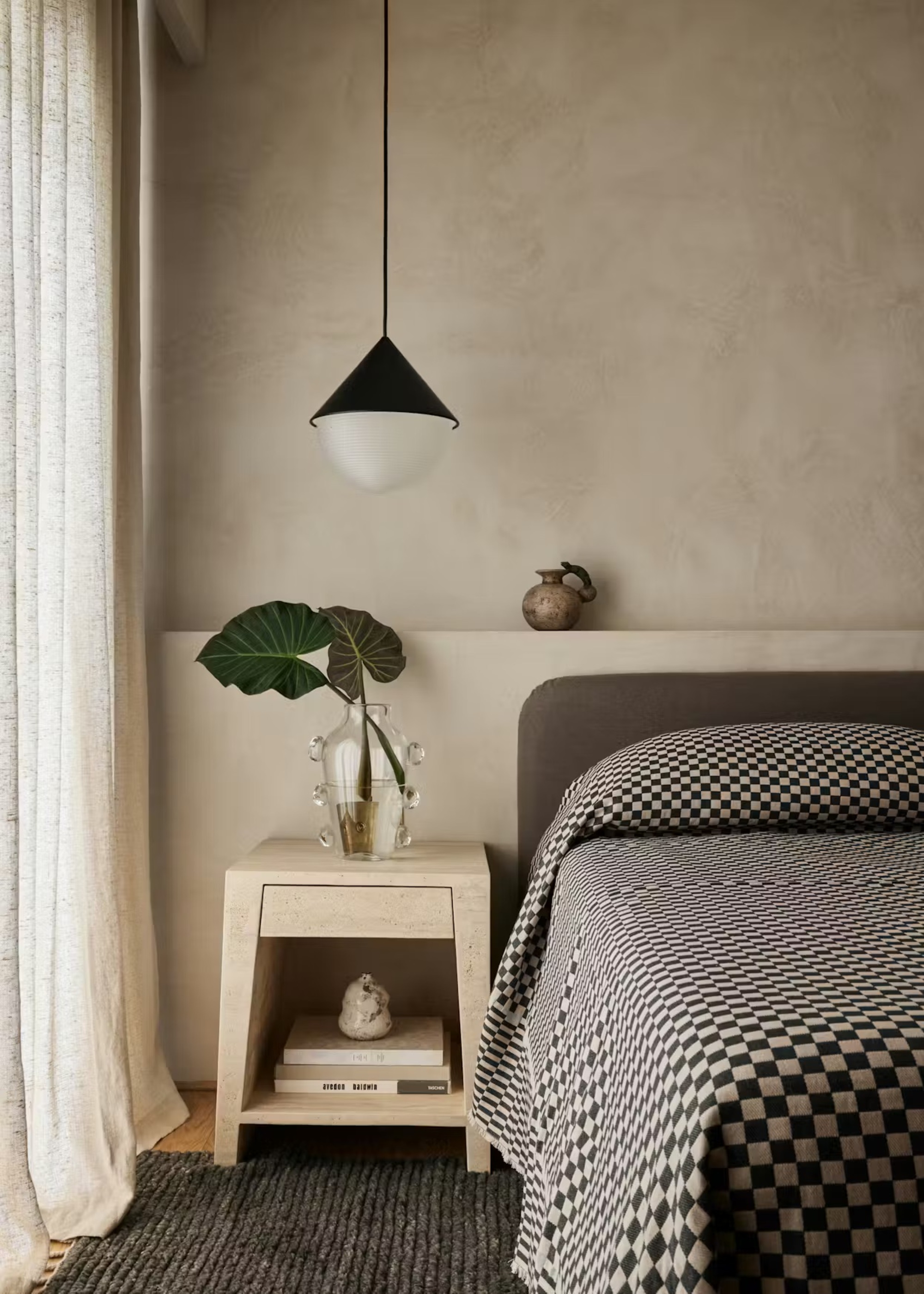 bedroom with plaster walls with niche behind the bed, side table with glass vase with branches, and gray headboard with checkered bedcover