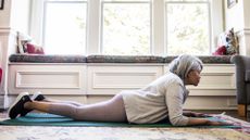 older woman lying on her front and forearms on an exercise mat in a living room setting with big windows behind her