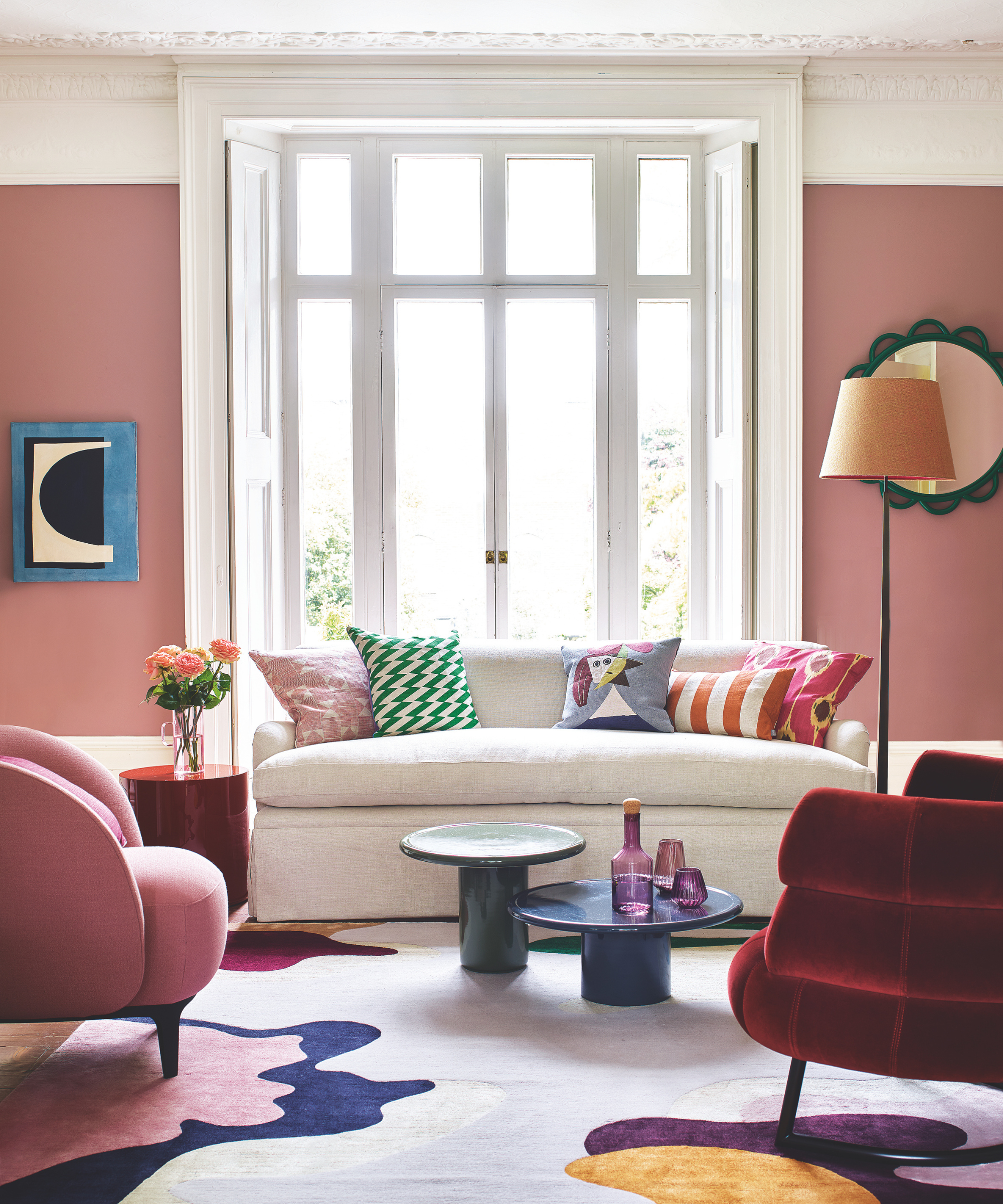 a colorful living room with pink walls, a white sofa with colorful pillows, a pink accent chair, a red accent chair, and a colorful rug