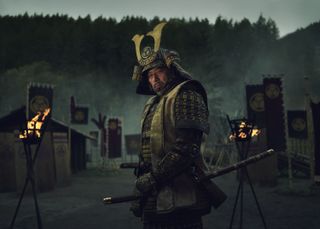Lord Yoshii Toranaga (Hiroyuki Sanada) stands in a small village in full armour. There are banners and lit torches behind him.