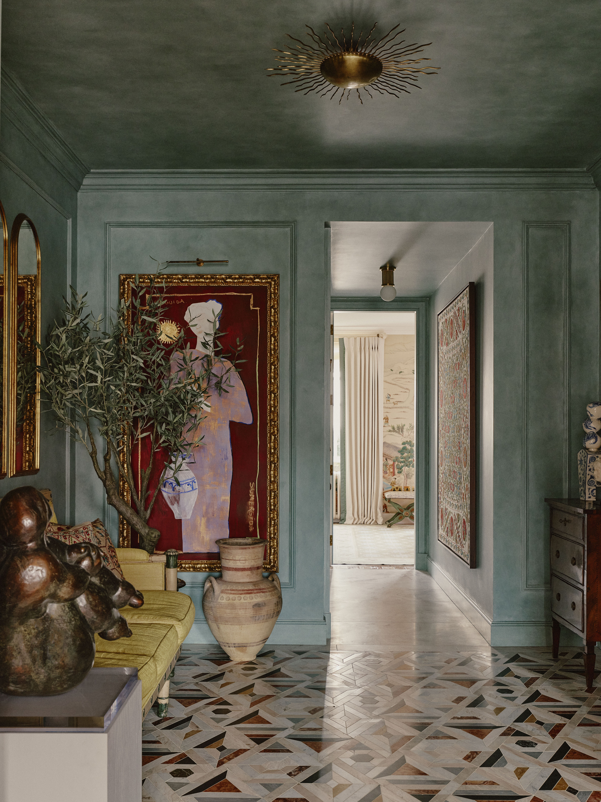 colorful entryway with green walls with molding, art in gilded gold frames, yellow sofa, plinth with sculpture, and mosaic-style tiled floor