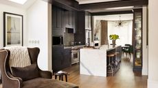 Modern kitchen and dining area with marble kitchen island, high stools, large brown suede armchair, dark cabinets and white walls.