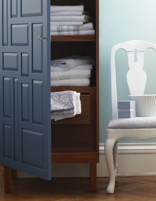 Bathroom cabinet with blue door open to piles of towels and linens with side chair alongside and wood floor