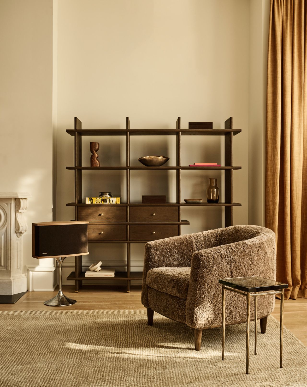 A beige living room with a fuzzy light brown accent chair, a metal side table with a glass top, a beige rug, and a large wooden shelving unit with an open back.